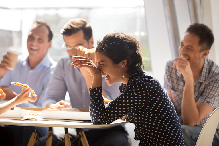 Woman and coworkers laugh at joke sitting around table with pizza