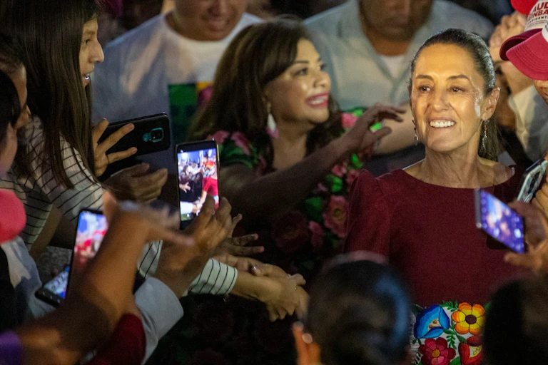 Claudia Sheinbaum smiling as she is embraced by a crowd of joyous people.