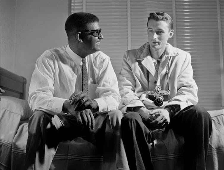 Smiling Black man wearing a tie sits on a bed while talking to a young white man with a camera hanging from his neck.