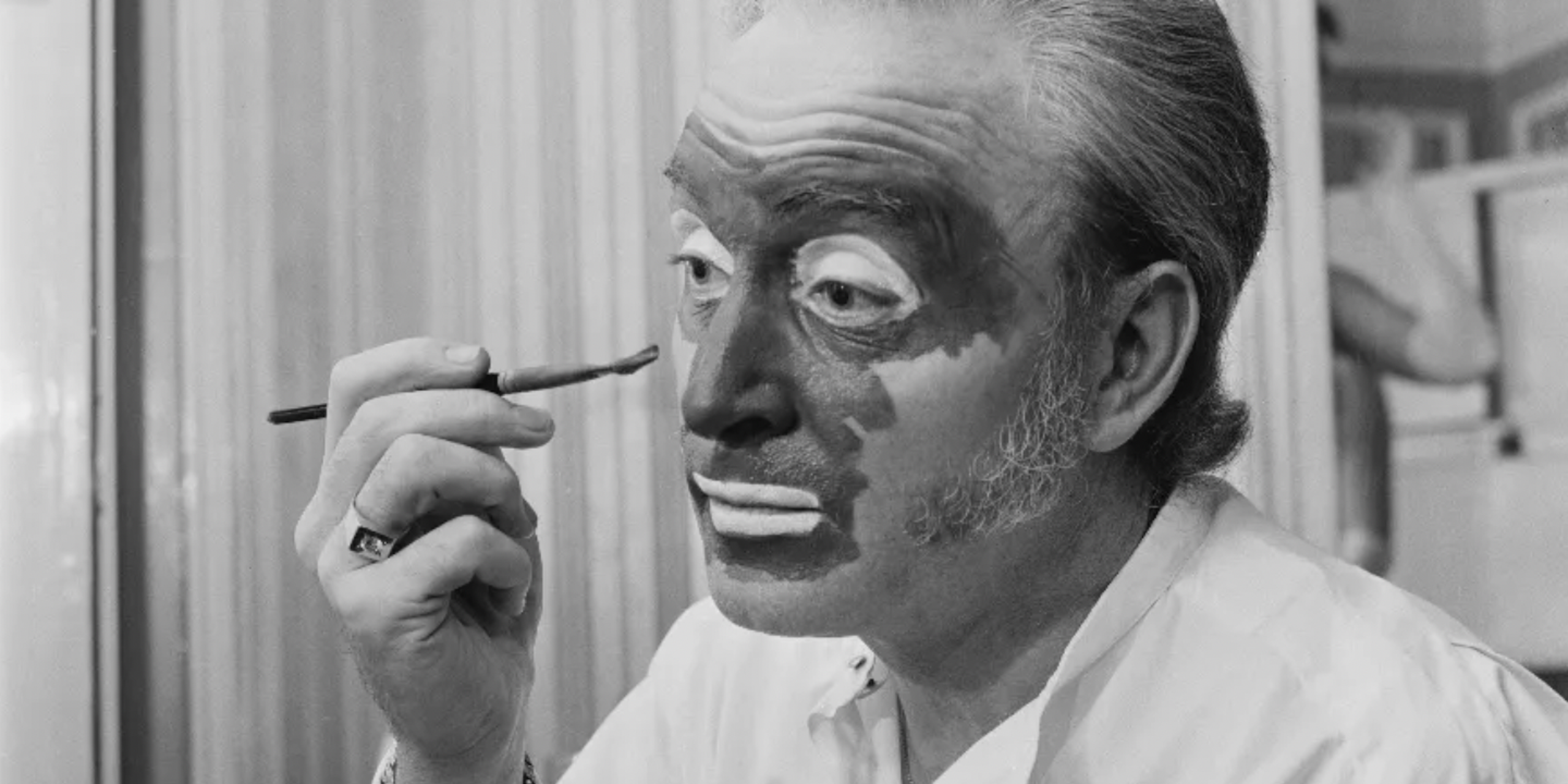 Black and white photo of white man applying dark paint to his face with a brush.