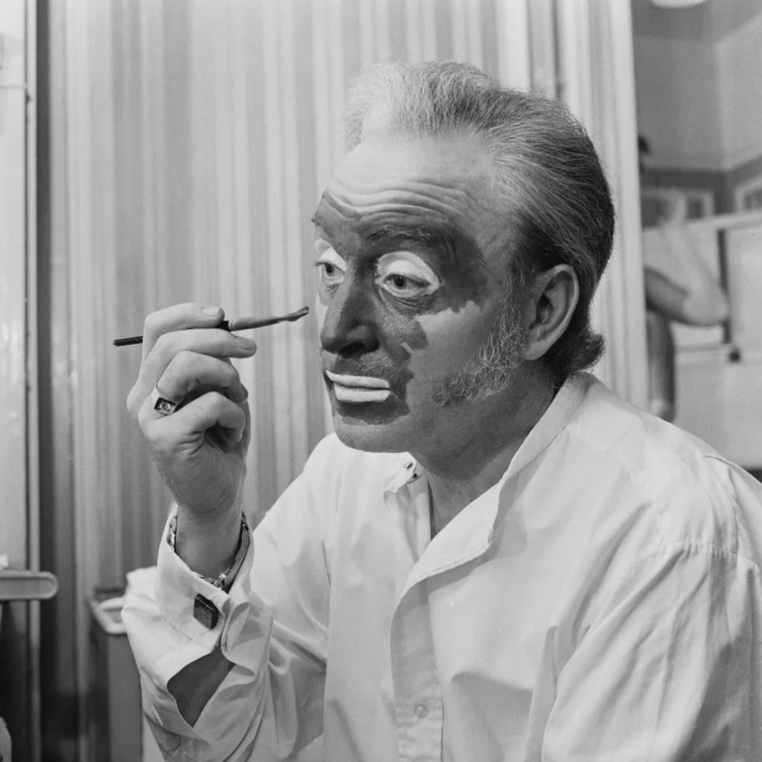Black and white photo of white man applying dark paint to his face with a brush.