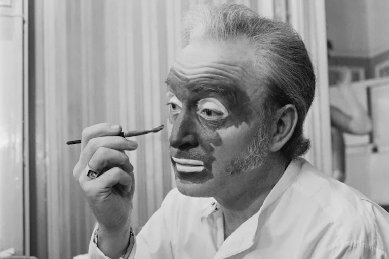 Black and white photo of white man applying dark paint to his face with a brush.