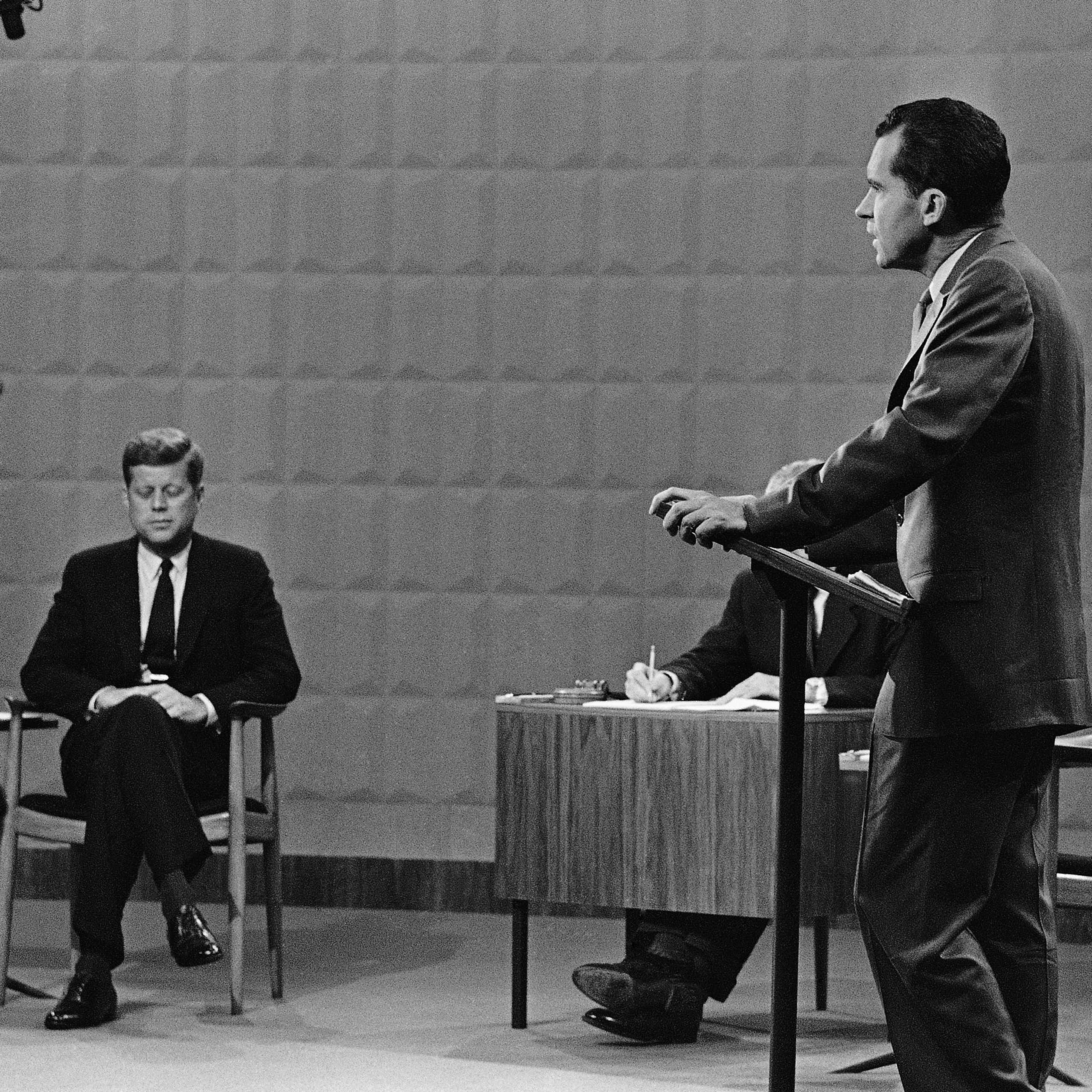 A black and white photo shows a man sitting down in a dark suit while another man stands at a lectern nearby him.