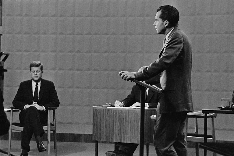 A black and white photo shows a man sitting down in a dark suit while another man stands at a lectern nearby him.