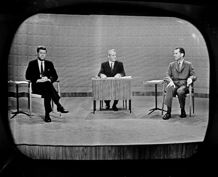 Three men are seen sitting in a black and white photo, which appears to be photo of a TV screen.