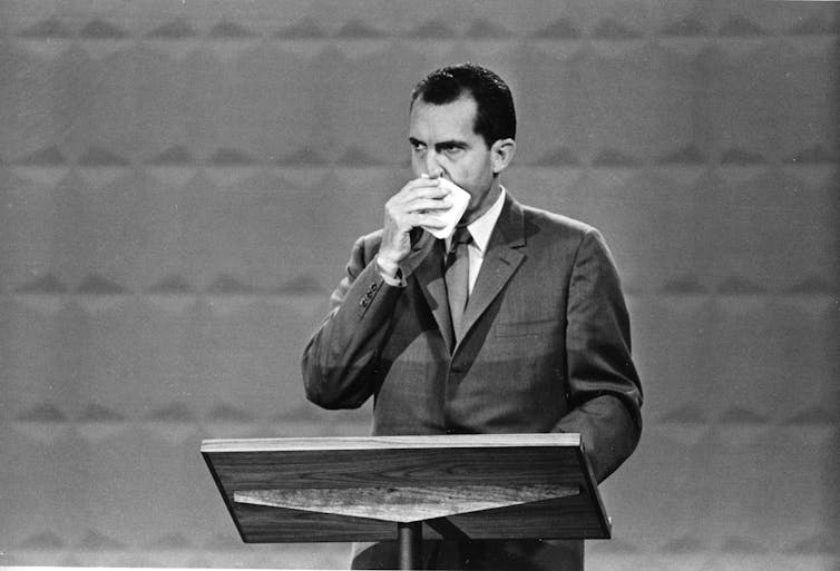 A black and white photo shows a man standing at a lectern, with a handkerchief covering his mouth.