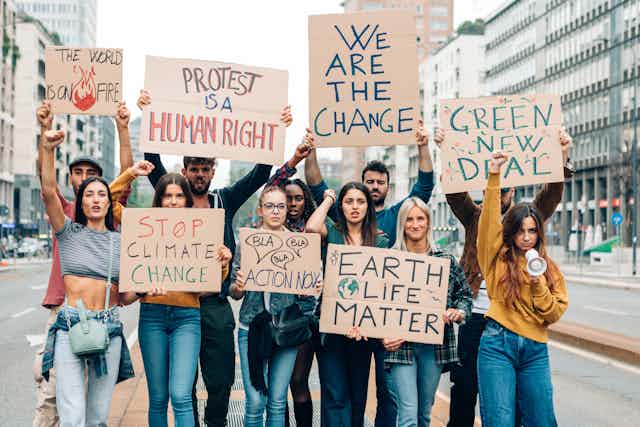group of 11 young people walking towards camera holding cardboard climate protest signs up above their heads, city in background, messages include 'we are the change' and 'stop climate change'