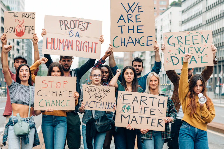 group of 11 young people walking towards camera holding cardboard climate protest signs up above their heads, city in background, messages include 'we are the change' and 'stop climate change'