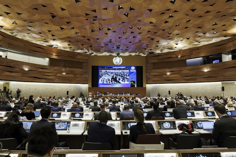 A large meeting room with rows of people sitting at screens with a panel of people at the front and a large viewing screen
