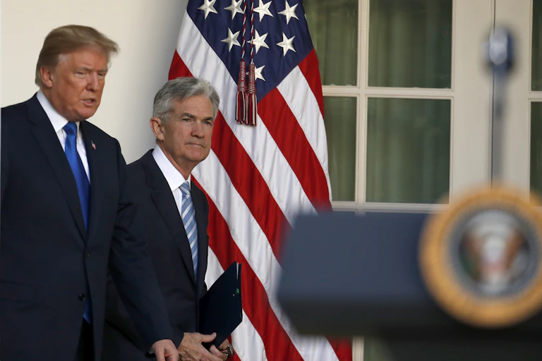Two men in dark suits walk to a platform with an American flag behind them