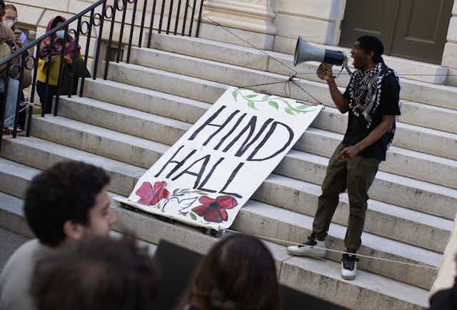 A person seen with a megaphone and a banner that says 'Hind Hall.'