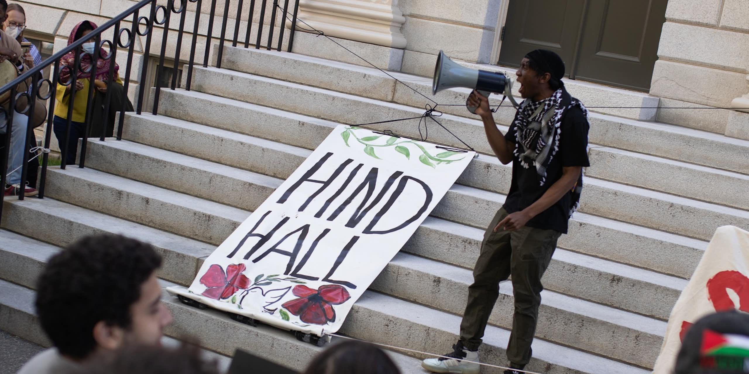 A person seen with a megaphone and a banner that says 'Hind Hall.'