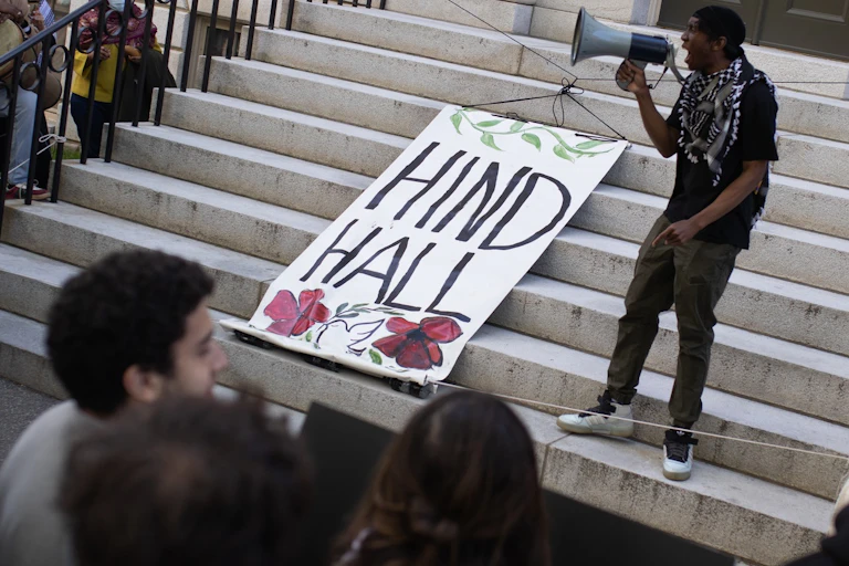 A person seen with a megaphone and a banner that says 'Hind Hall.'