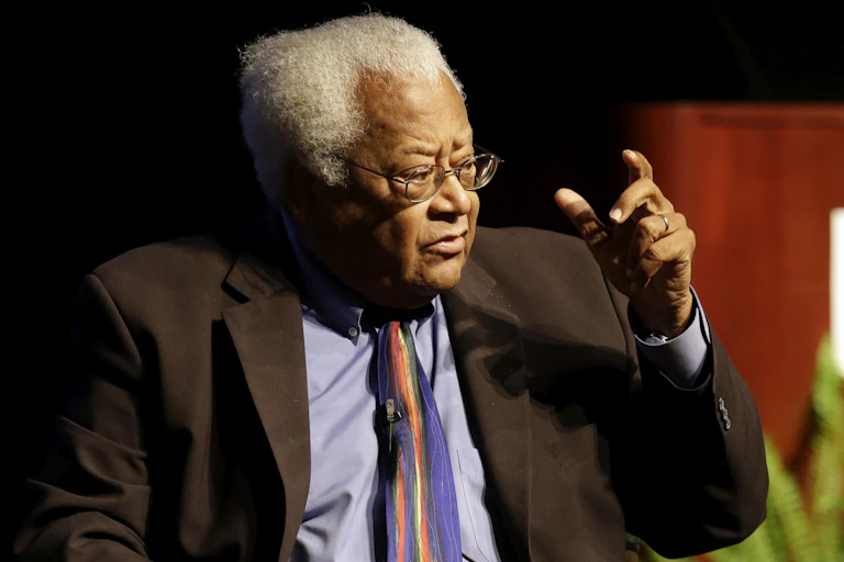 A gray-haired, bespectacled Black man, wearing a brown suit and a tie, gesticulates with his hand as he speaks.