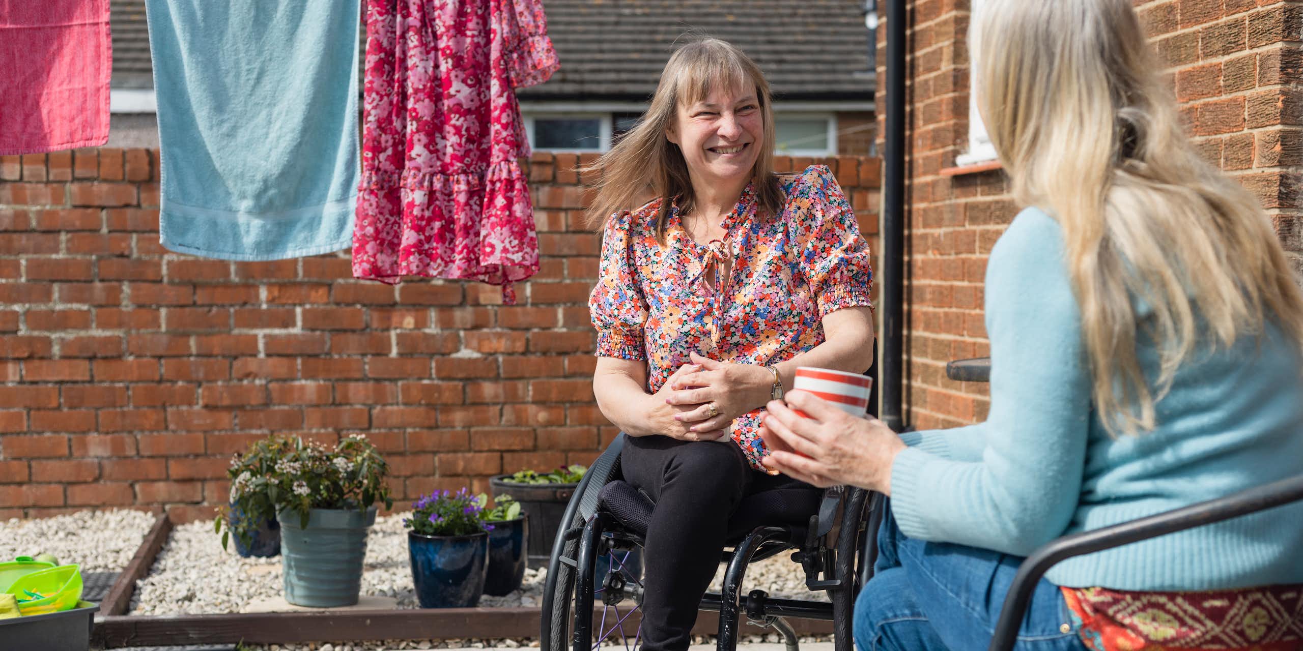 two women talking in home courtyard with washing hanging nearby. One woman using wheelchair