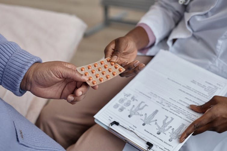 Health worker handing blister pack of medicine to another, clipboard in hand