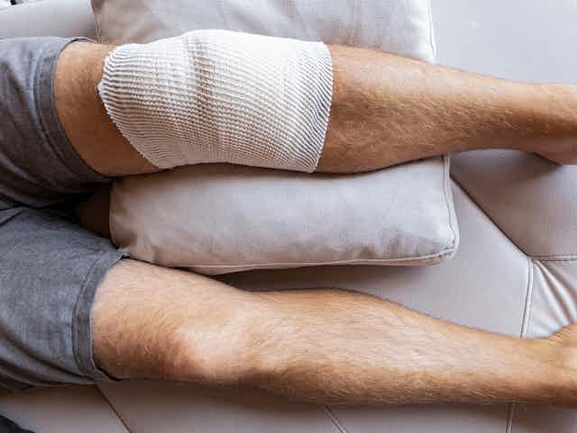 Man lying on sofa, legs stretched out, one knee bandaged after surgery