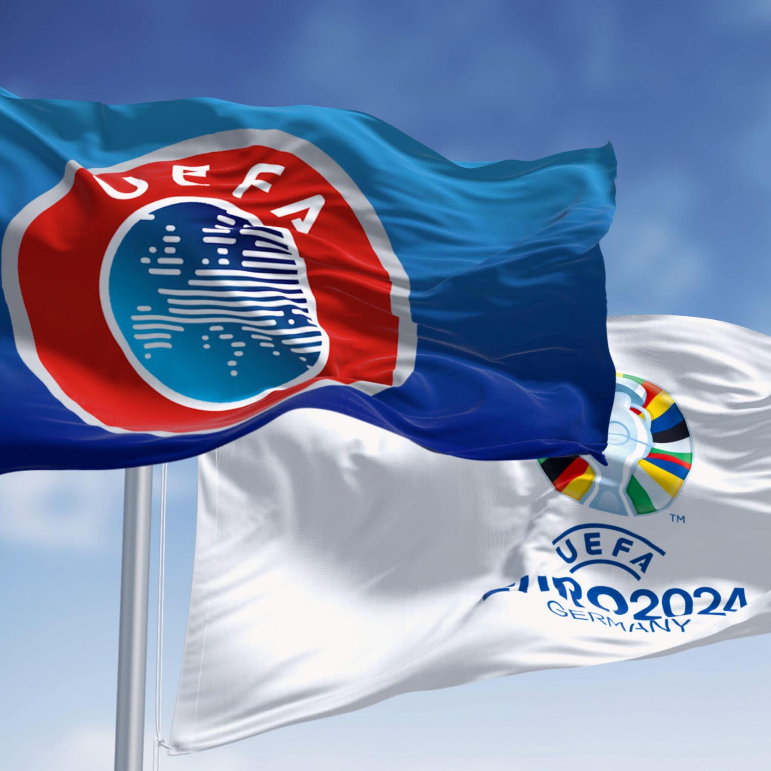 A Uefa flag and a Euro 2024 flag flying against a blue sky.