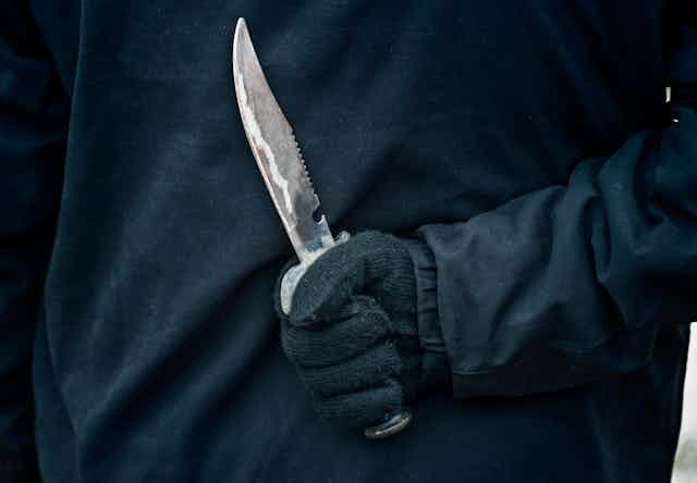A gloved hand holding a sharp knife behind the user's back