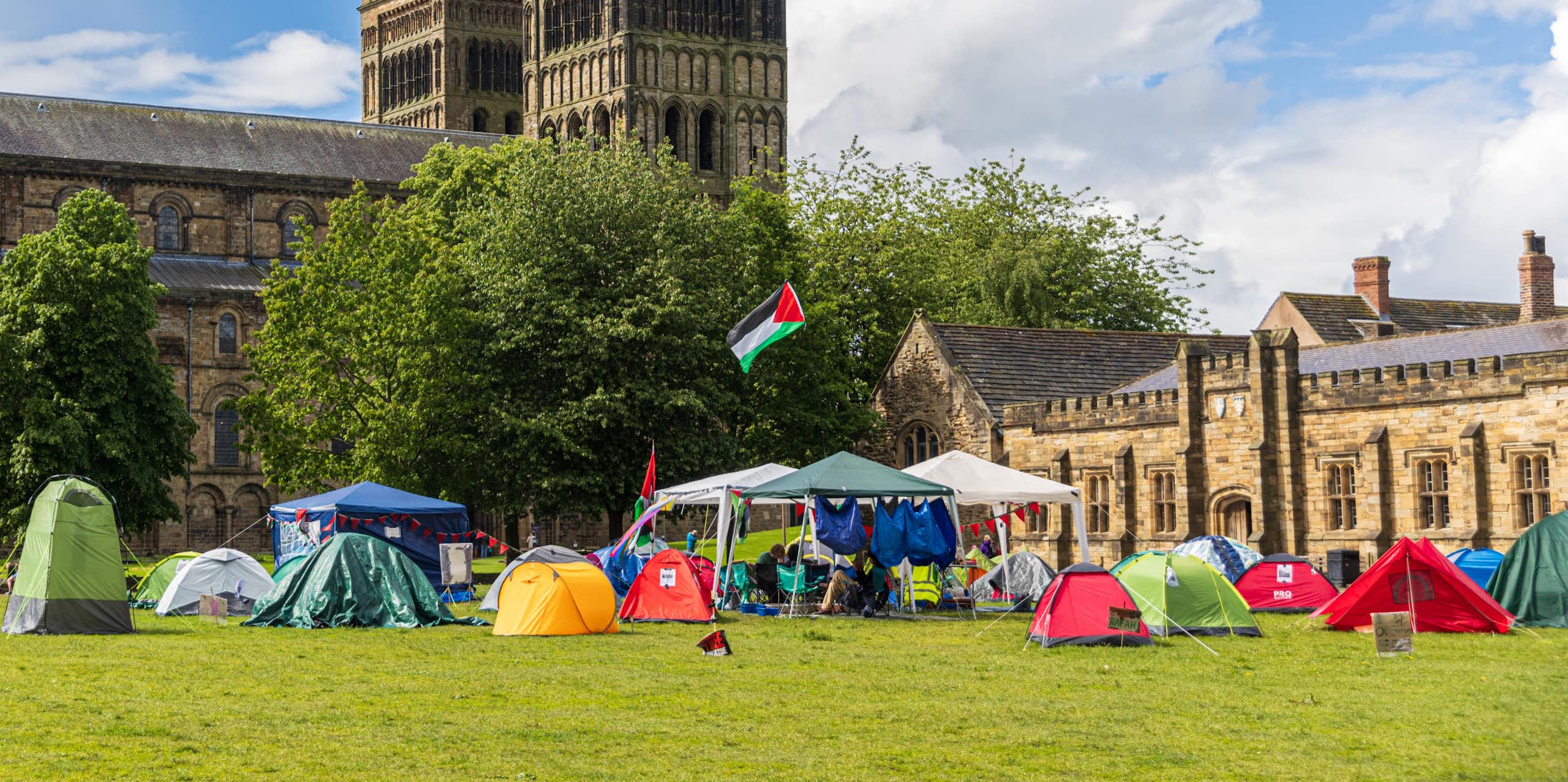 A small group of tents with a flying Palestinian flag on a green in front of a large gothic cathedral