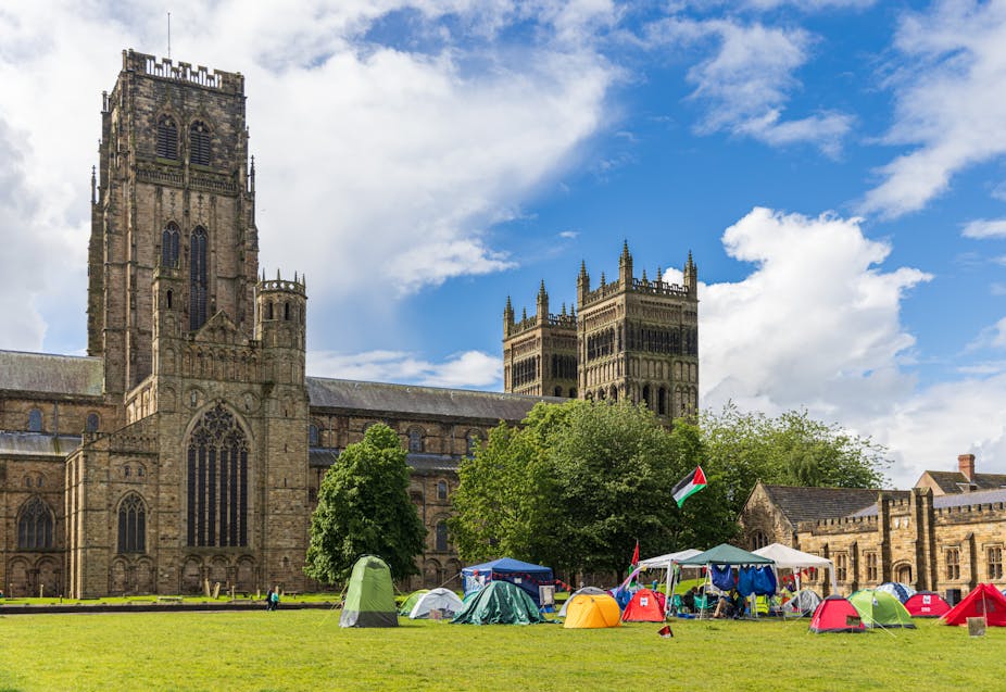A small group of tents with a flying Palestinian flag on a green in front of a large gothic cathedral