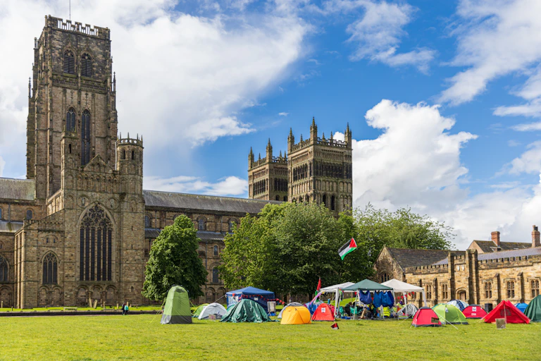A small group of tents with a flying Palestinian flag on a green in front of a large gothic cathedral