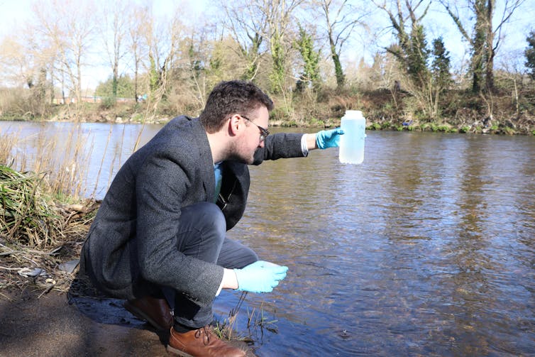 A man crouches on the side of a river wearing blue surgical gloves holding a container of water.