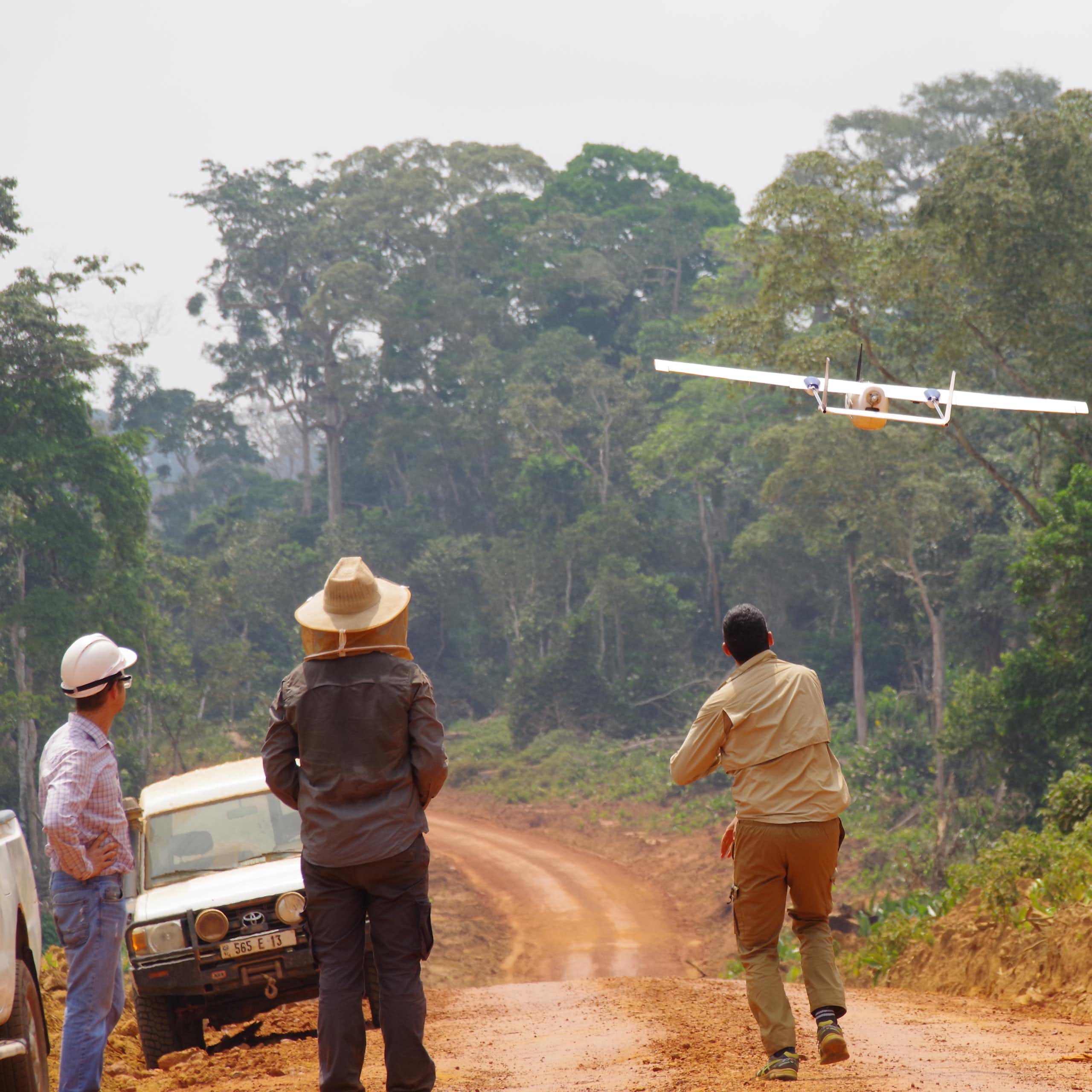 trois hommes derrière un drone qui décolle au-dessus d'une forêt