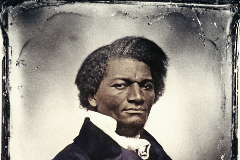 A Black man wearing a dark suit and white shirt sits behind a desk.