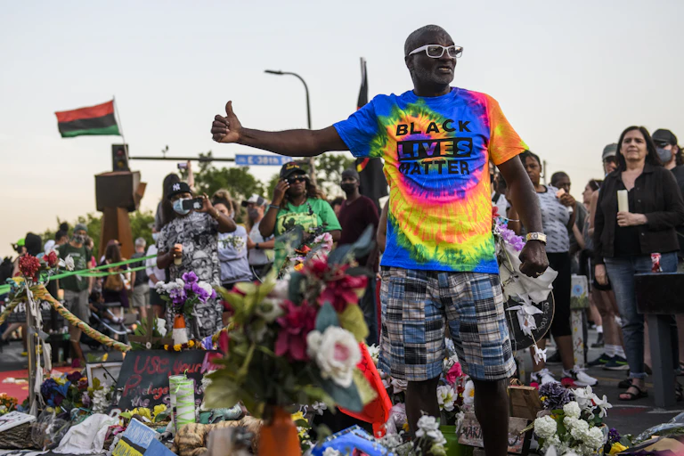 A man in a Black Lives Matter t-shirt talks to a large crowd outside.