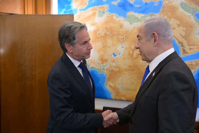 US secretary of state, Antony Blnken, shakes hands with the Israeli prime minister in front of a world map.