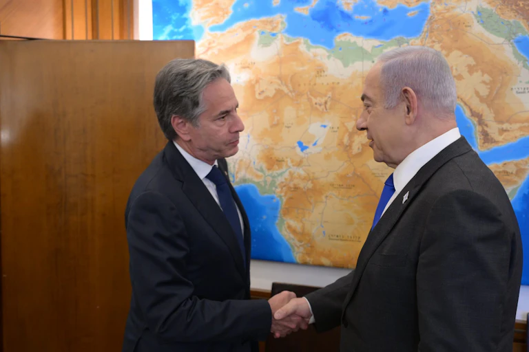US secretary of state, Antony Blnken, shakes hands with the Israeli prime minister in front of a world map.