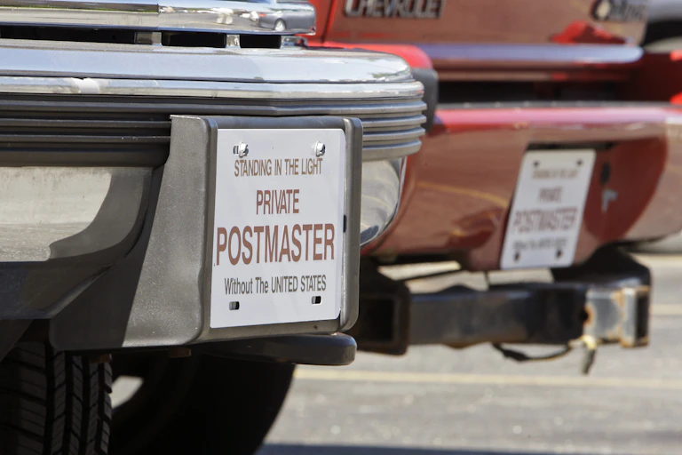 A close-up of a car with a sign in place of a license plate reading, in part 'Private postmaster without the United States.'