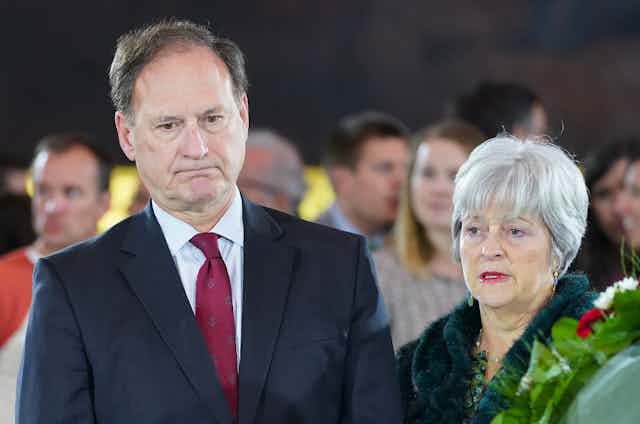 a man in a business suit and a woman wearing a coat stand beside each other with a group of people behind them