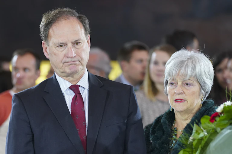 a man in a business suit and a woman wearing a coat stand beside each other with a group of people behind them