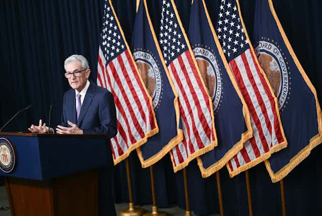 A central banker in a navy blue suit stands behind a podium and in front of a series of American flags. He appears to be speaking, and is gesticulating for emphasis.