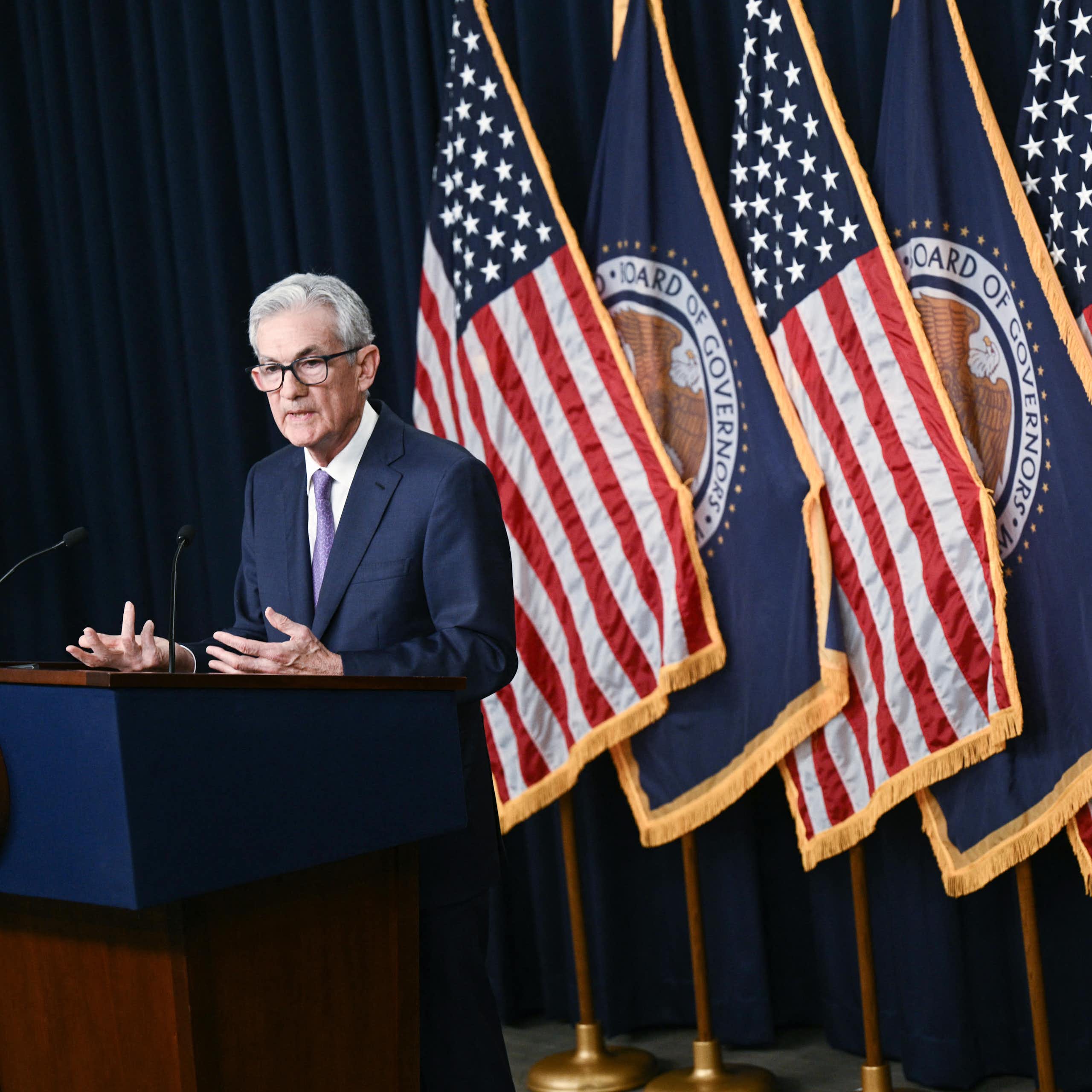 A central banker in a navy blue suit stands behind a podium and in front of a series of American flags. He appears to be speaking, and is gesticulating for emphasis.