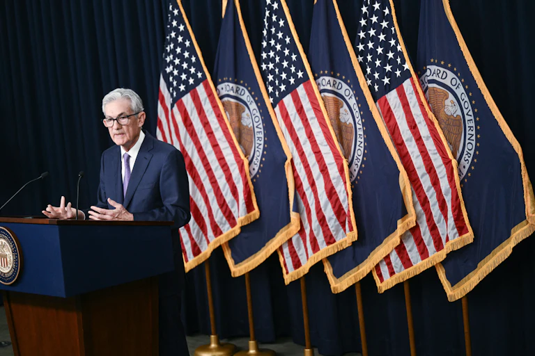 A central banker in a navy blue suit stands behind a podium and in front of a series of American flags. He appears to be speaking, and is gesticulating for emphasis.