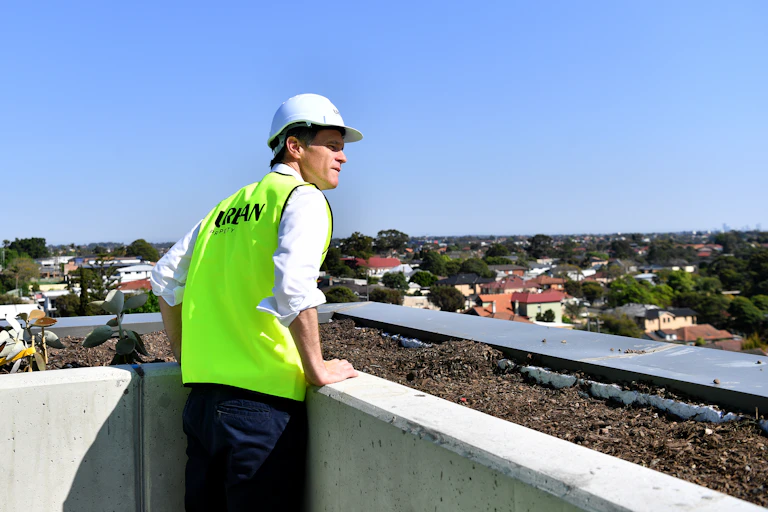 A man in a hard hat and hi-viz vest looks out over an apartment balcony railing