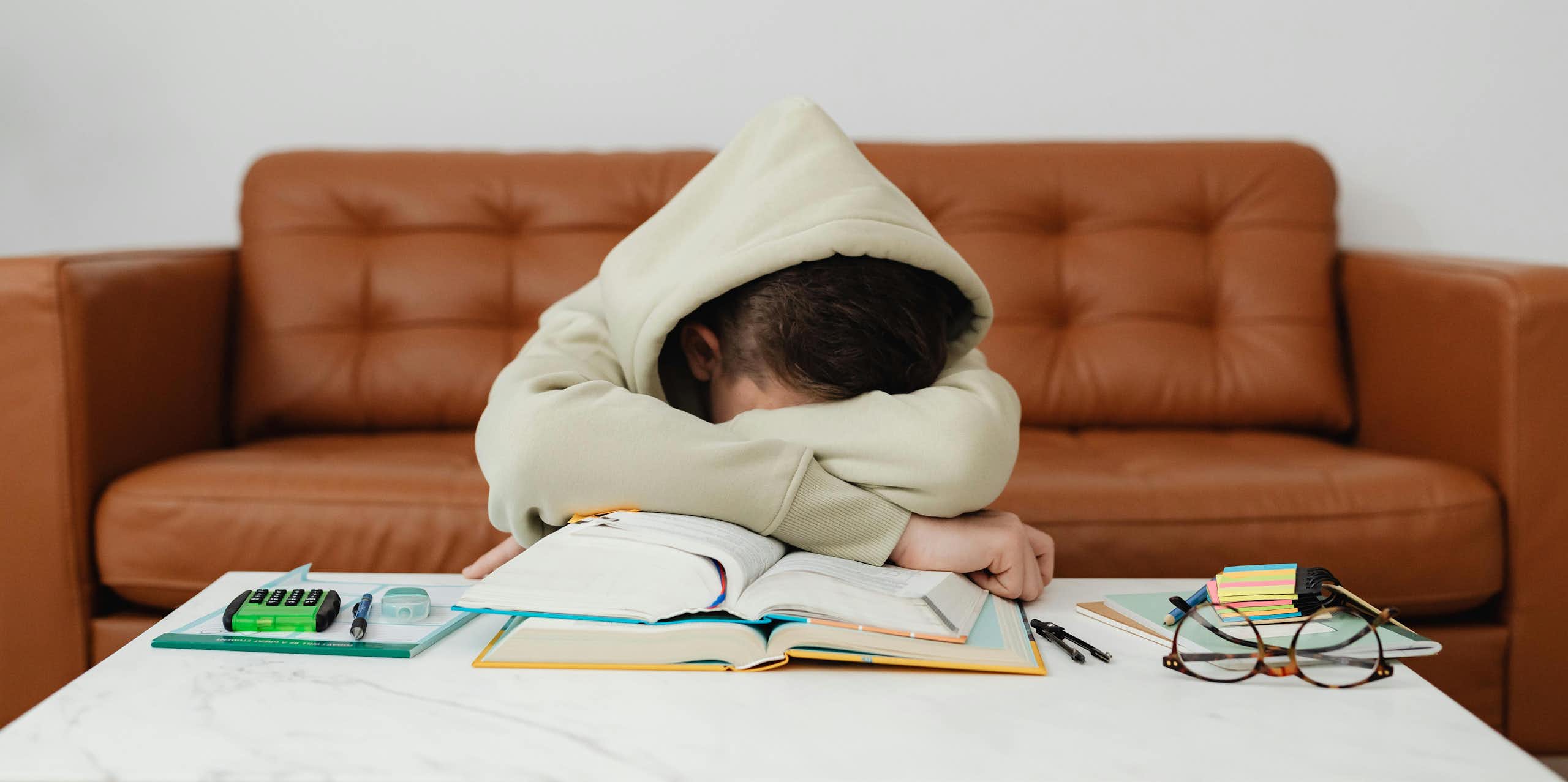 A young person in a hoodie rests their head in the arms on top of books. 