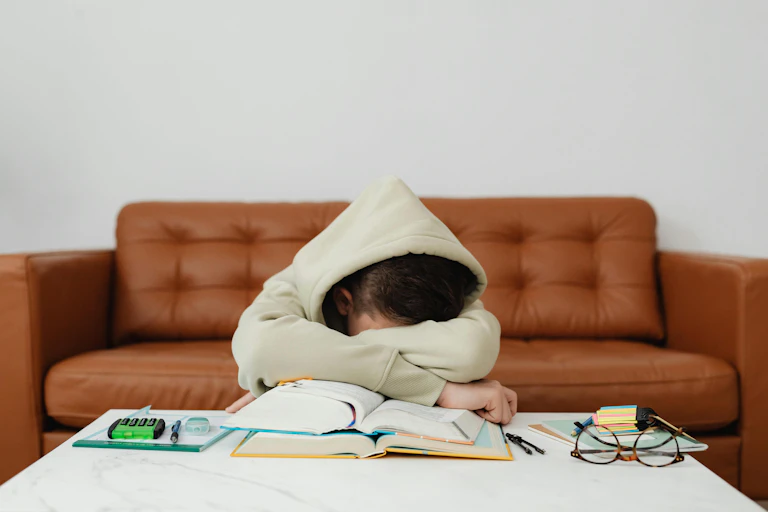 A young person in a hoodie rests their head in the arms on top of books.
