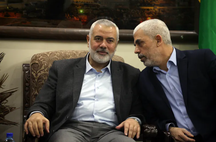 Two middle-aged men sit talking, one is saying something into the other's ear.