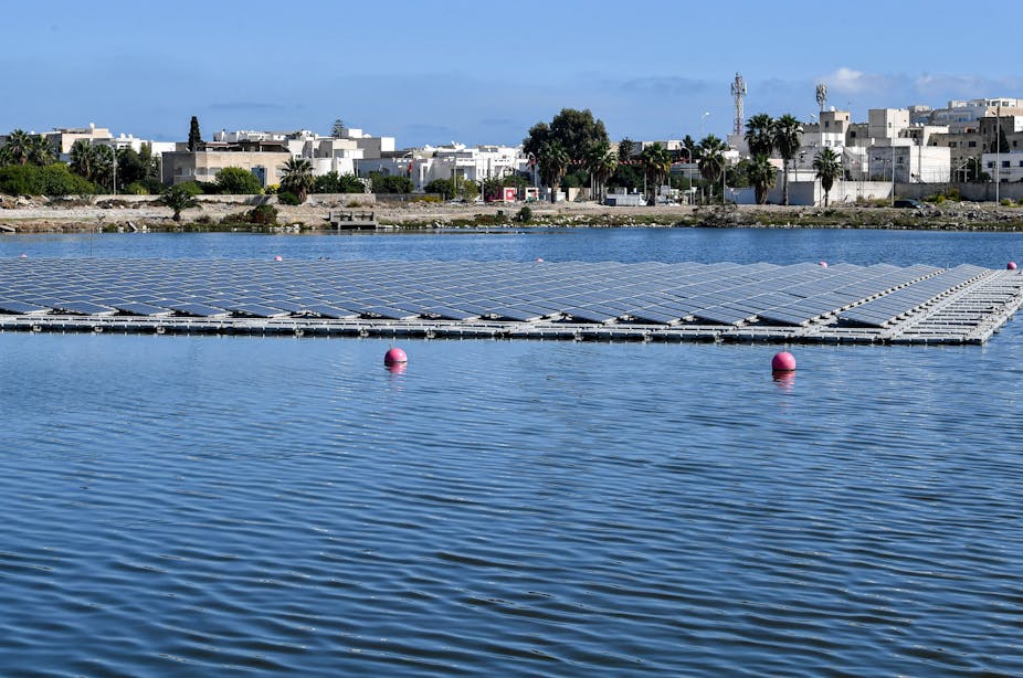 Dozens of lines of solar panels float on a lake in front of a Tunisian scene of traditional whitewashed buildings
