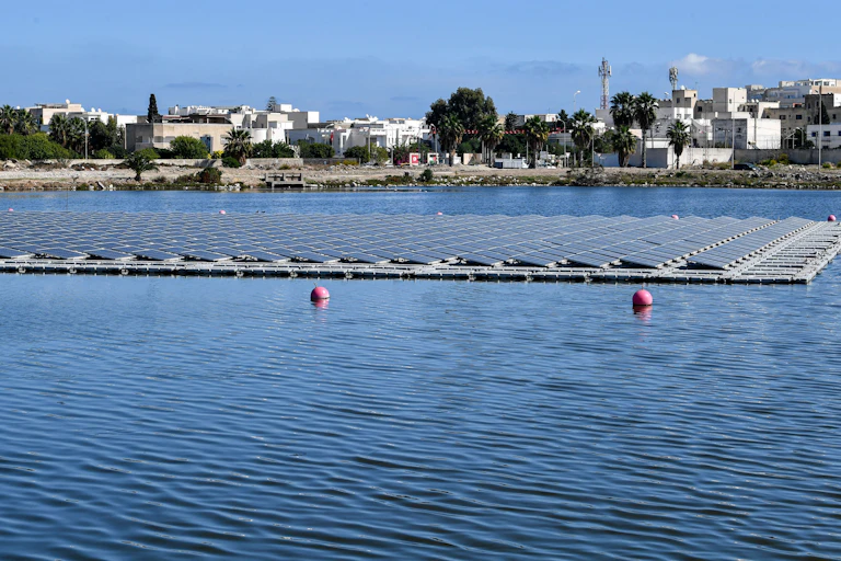 Dozens of lines of solar panels float on a lake in front of a Tunisian scene of traditional whitewashed buildings