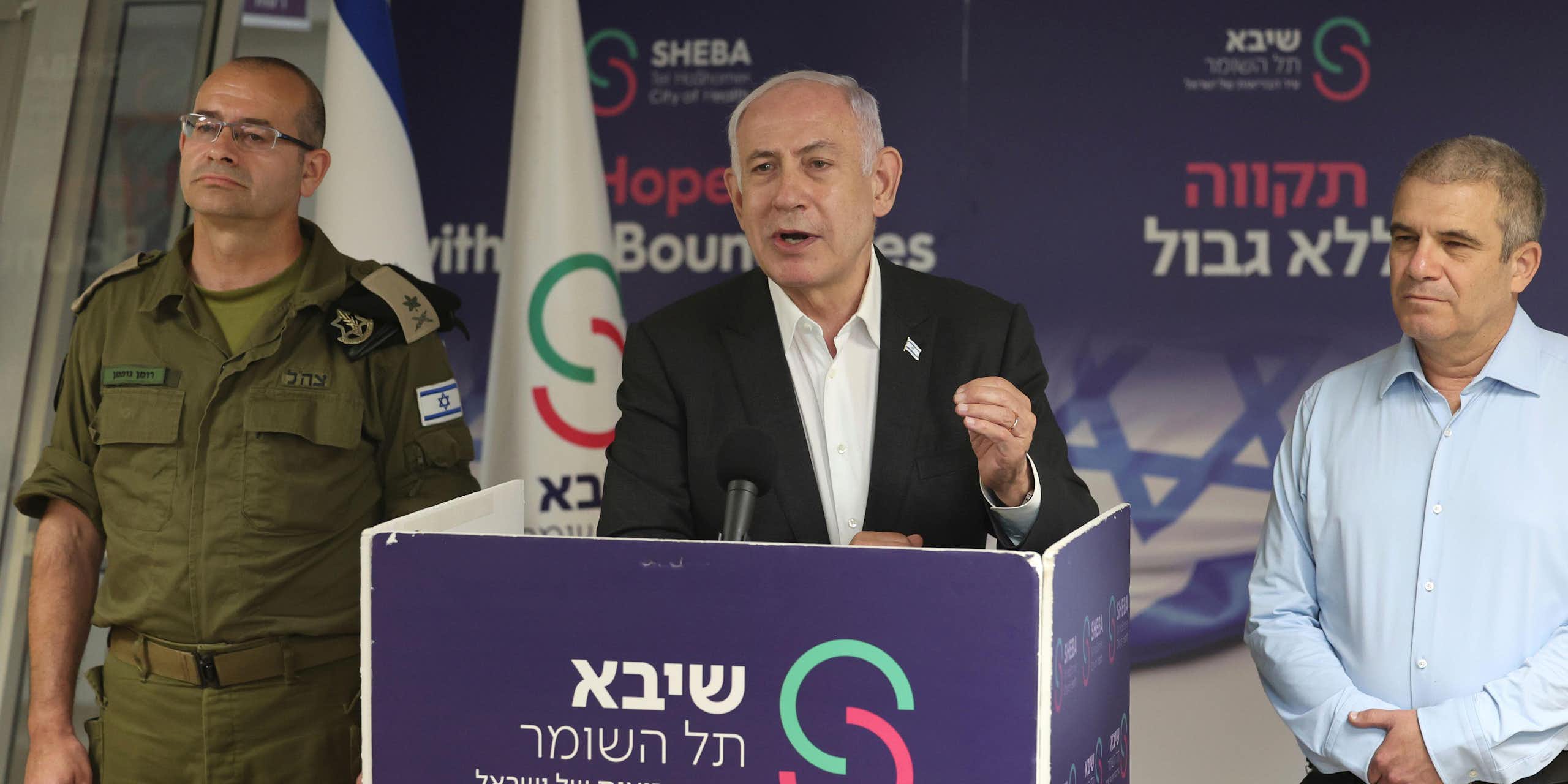 Israeli prime minister Benjamin Netanyahu delivers a speech from a podium, flanked by a military and a political aide.