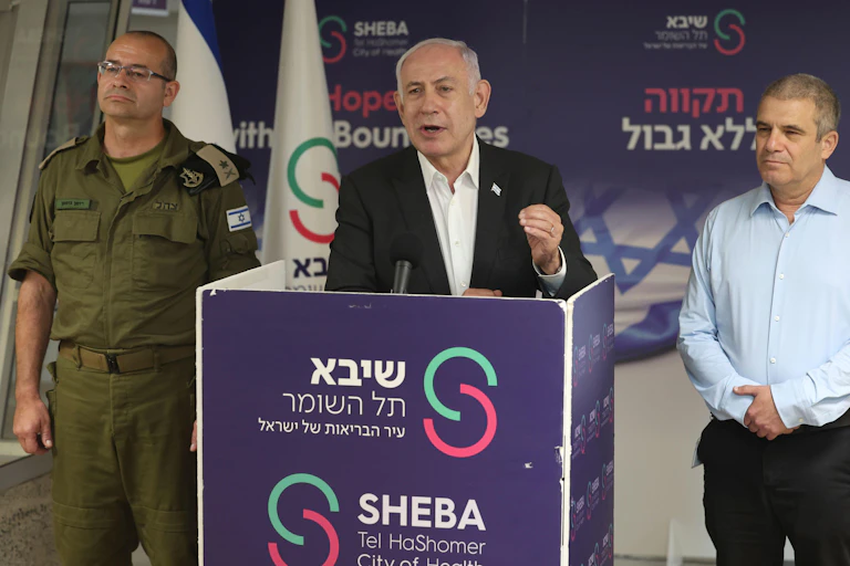 Israeli prime minister Benjamin Netanyahu delivers a speech from a podium, flanked by a military and a political aide.