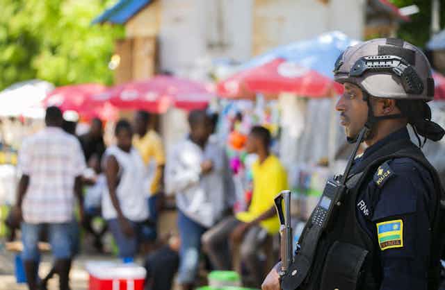 A man staring straight ahead wearing military gear and holding a gun, with a blue, yellow and green flag on his left arm. There are a group of people going about their business blurred out in the background
