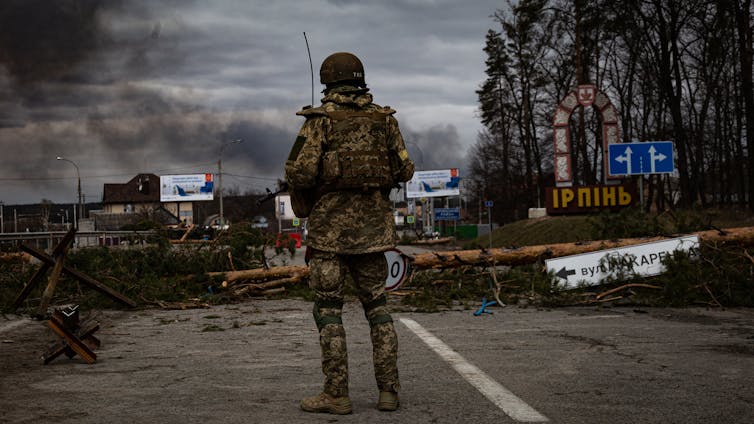 A soldier standing on a damaged road as smoke rises in the background.