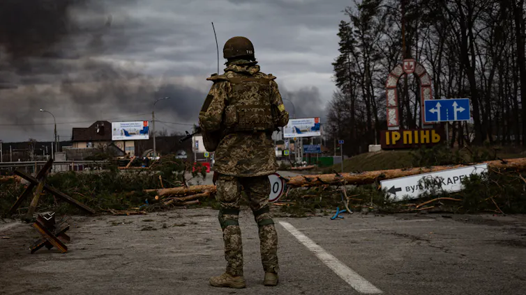 A soldier standing on a damaged road as smoke rises in the background.