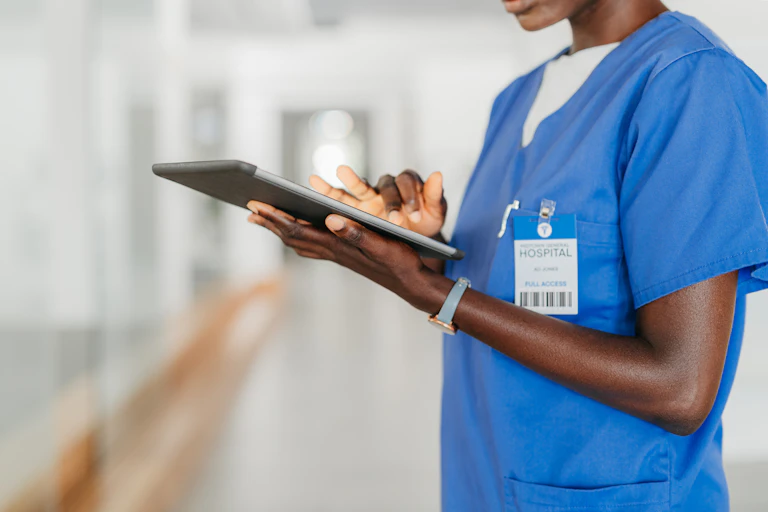Woman wearing blue hospital scrubs and a hospital nametag, standing in a corridor and holding a tablet computer